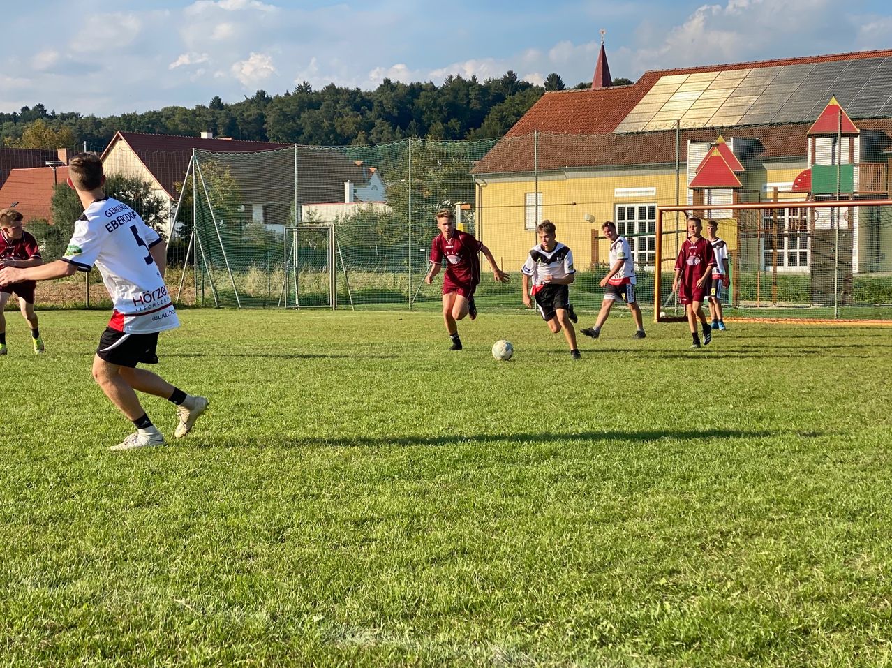 Eine Gruppe von Fußballspielern läuft auf einem Feld mit einem Fußball in der Mitte. Im Hintergrund befinden sich Gebäude und Häuser mit Solarpanelen.