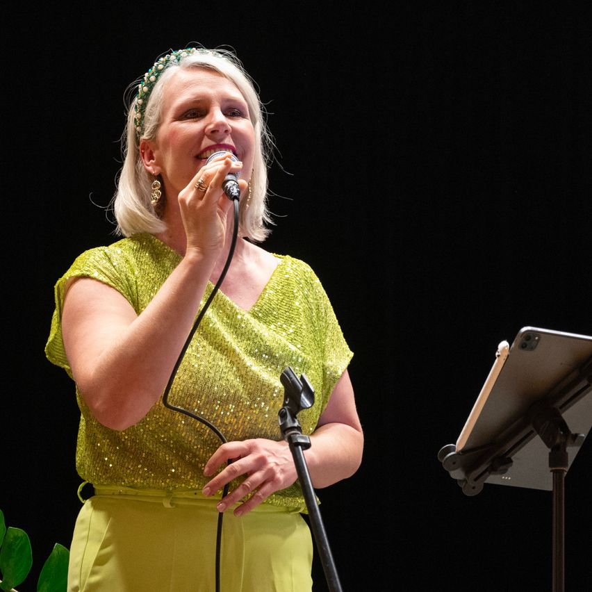 A woman in a sparkling green dress is singing into a microphone, smiling, with a plant on the left and a music stand on the right.