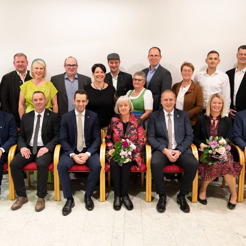 A group of people in formal attire are seated and standing in front of a white wall, smiling for a photograph. There are both men and women, all dressed in business suits, dresses, and coats. Some are holding flowers.