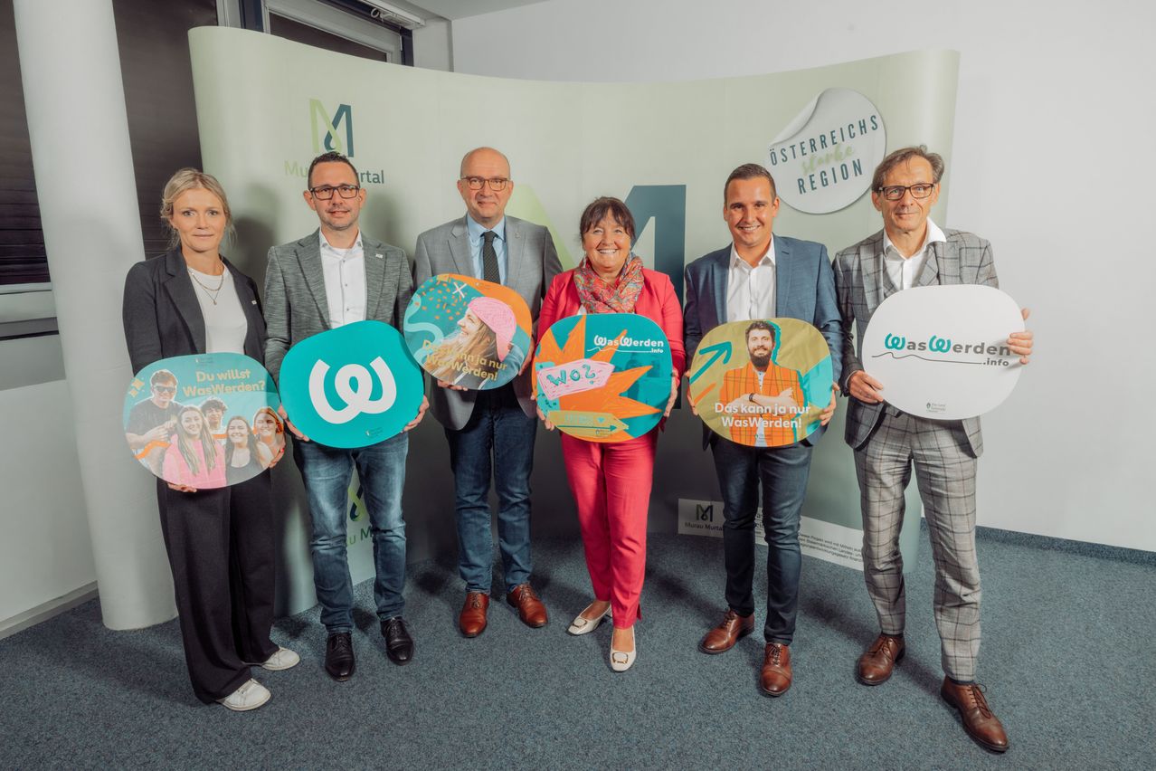 Sechs formell gekleidete Personen stehen vor einer Kulisse mit einem M-Logo und Text und halten Schilder zur Förderung von 'WasWerden'. Sie lächeln und scheinen sich bei einer Veranstaltung zu befinden.