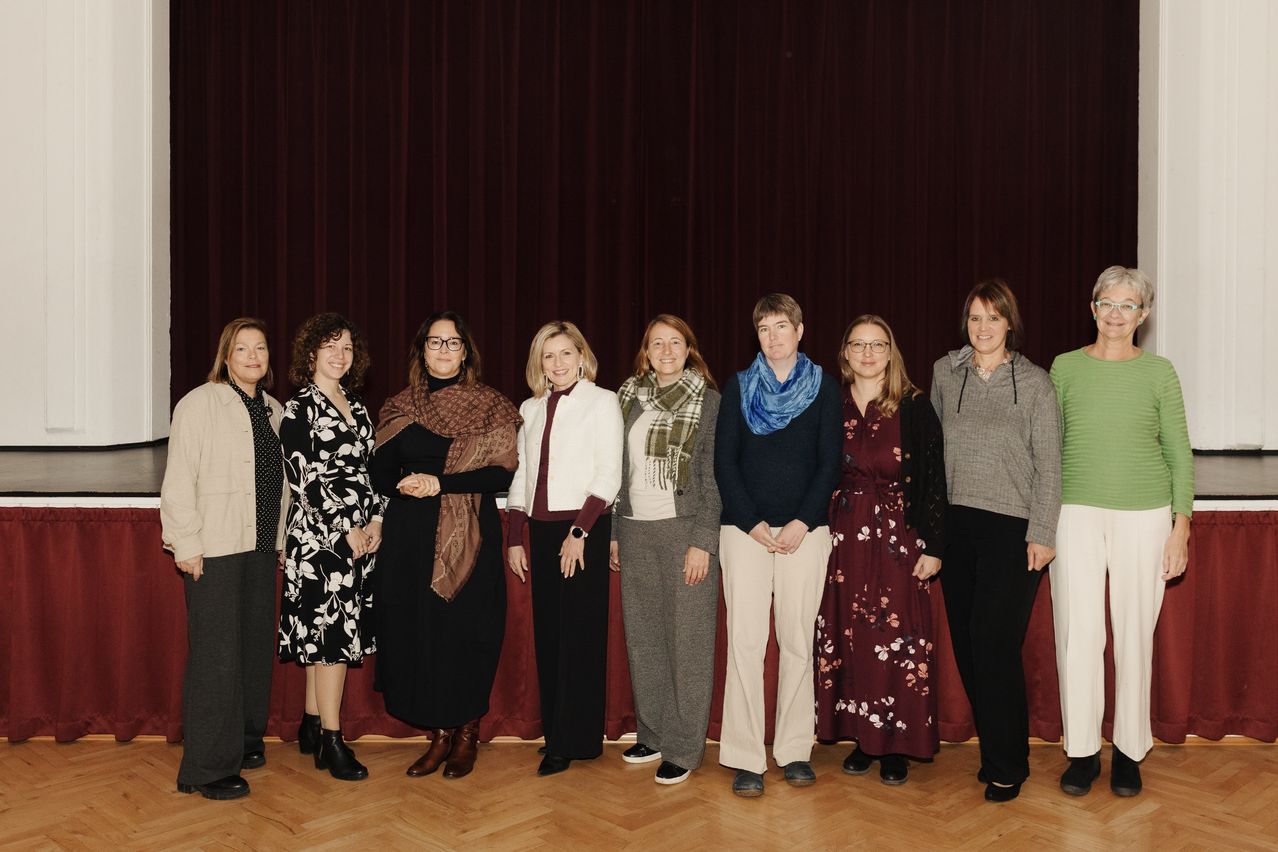 Eine Gruppe von Frauen in verschiedenen Winterkleidungen steht vor einer Bühne mit roten Vorhängen, möglicherweise bei einer Konferenz.