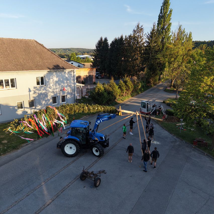 Eine Gruppe von Menschen hebt eine lange Holzbalken mit einem blauen Traktor. Der Traktor steht auf der Straße mit bunten Bändern, die daran hängen. Das Gebäude dahinter hat ein braunes Dach.