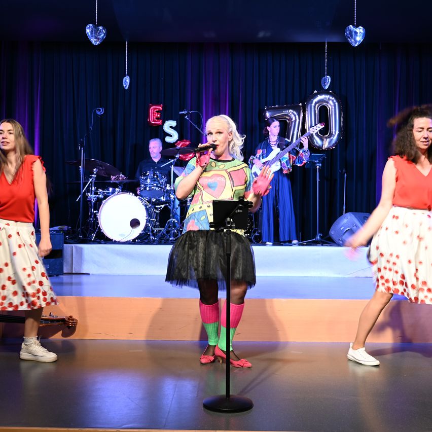 A woman sings on stage with a microphone while three women dance. Behind her, a band plays on stage with a drum set and a guitar.