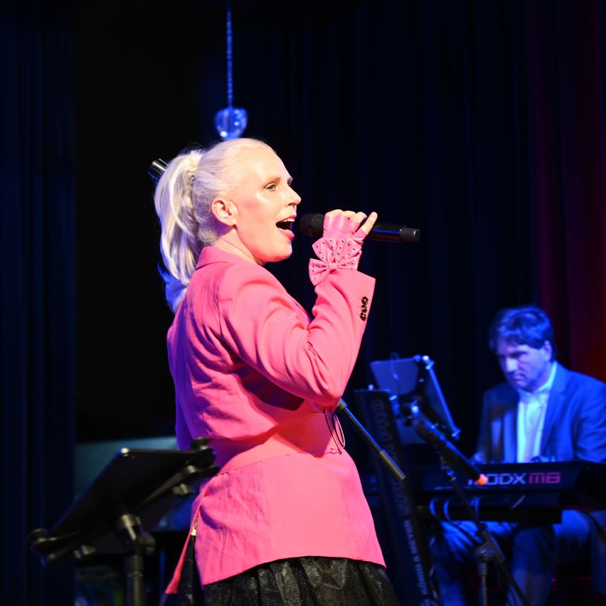 A woman in a pink blazer is singing on stage, holding a microphone. Behind her, a man plays a keyboard. The stage is illuminated with pink and blue lighting.