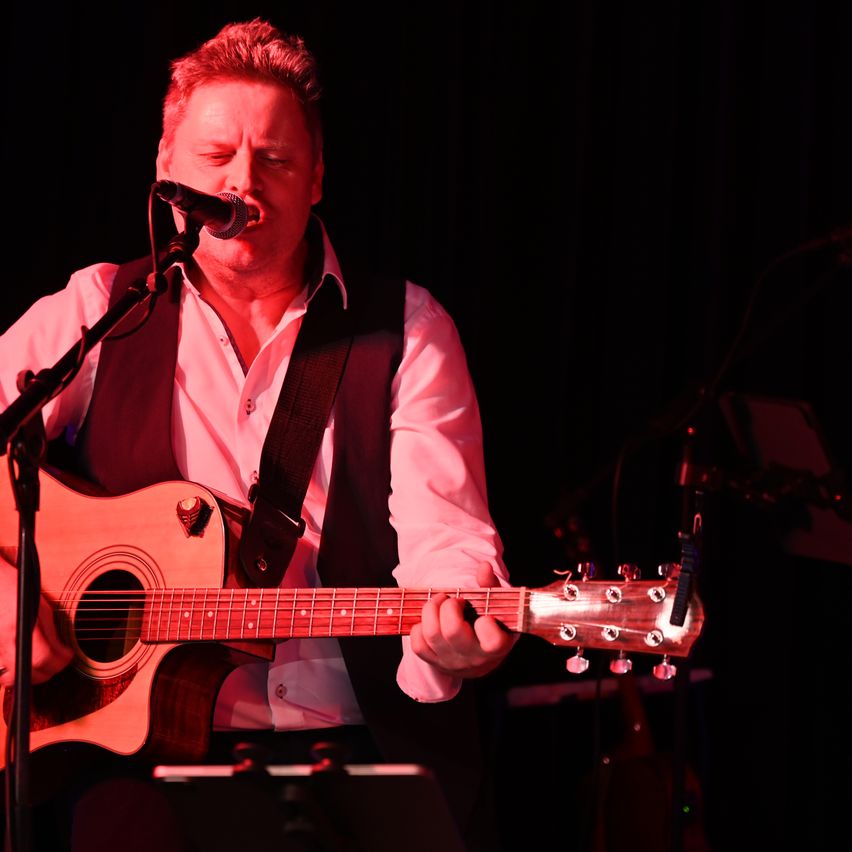 A man performs on stage with a guitar, singing into a microphone, illuminated by red lighting.