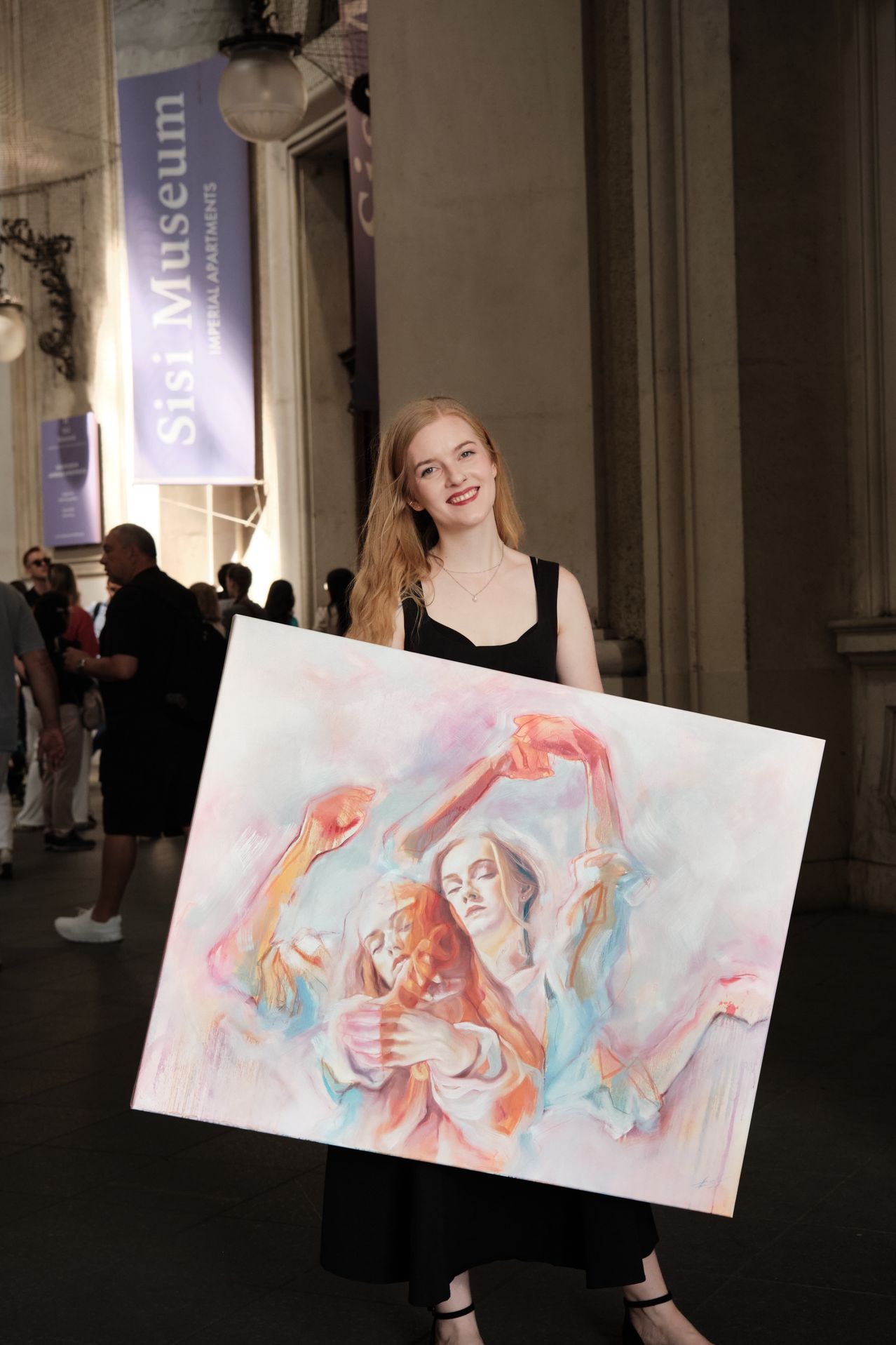 A woman holds a large painting in front of a crowd, possibly at an art exhibit. She smiles and looks at the camera. The painting depicts a woman in red, blue, and white.