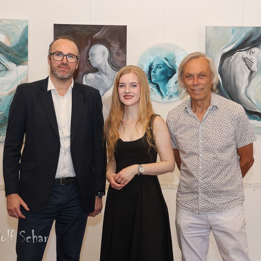 Three people stand in front of paintings, smiling. A man in a suit, a woman in a black dress, and an older man in a patterned shirt.