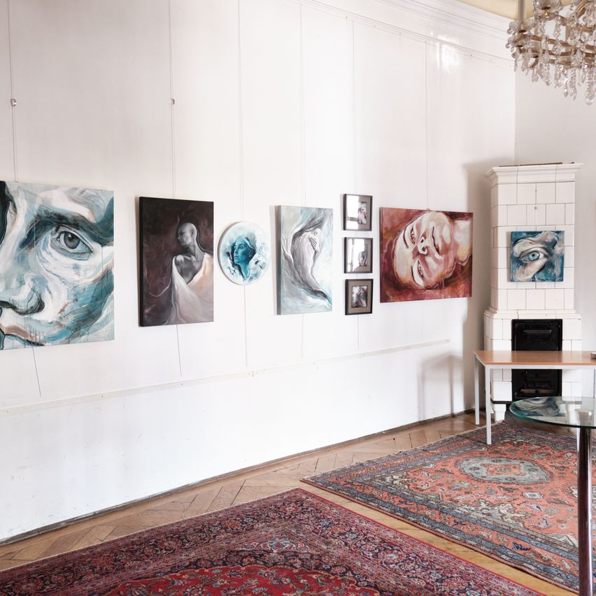 An art gallery space with white walls, featuring various paintings. A chandelier hangs from the ceiling. A rug covers part of the floor.