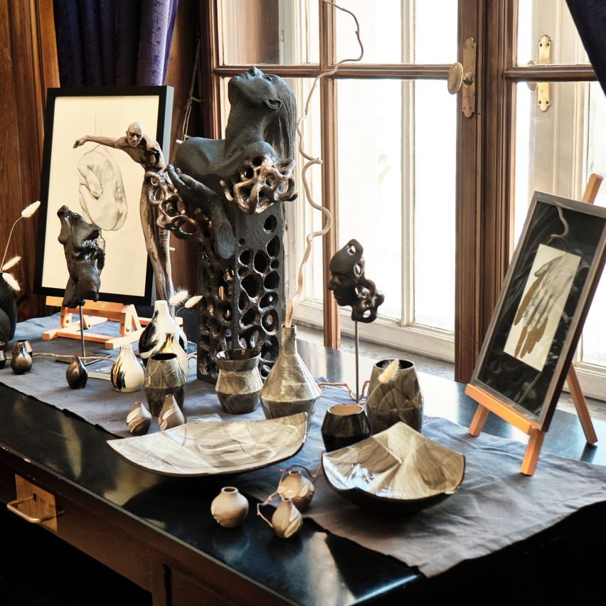 A table with several ceramic and clay sculptures on display, including a large abstract human figure, vases, and plates. A framed drawing is placed behind the sculptures, and a smaller framed piece is propped up on an easel.