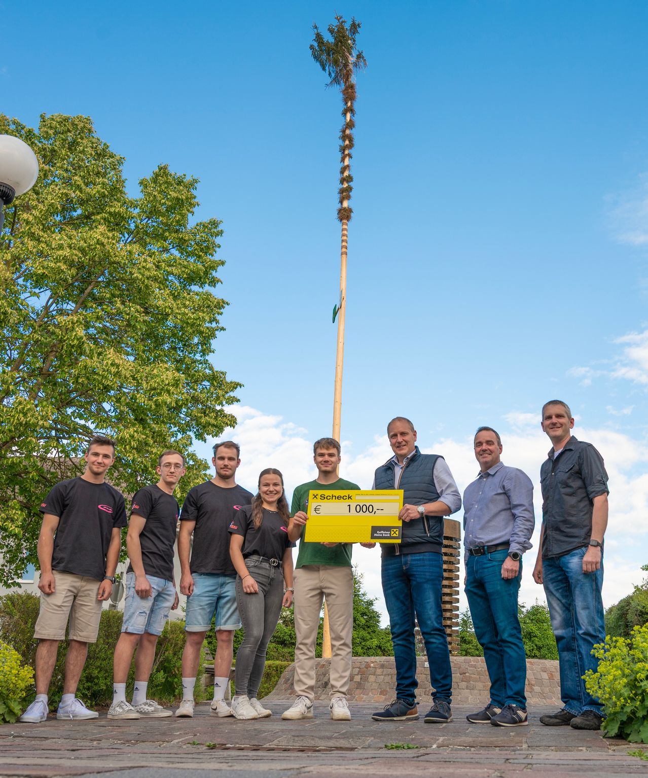 Eine Gruppe von Männern und Frauen steht im Freien unter blauem Himmel und hält einen großen gelben Scheck über 1.000 Euro. Sie scheinen zu feiern oder einen Preis entgegenzunehmen.