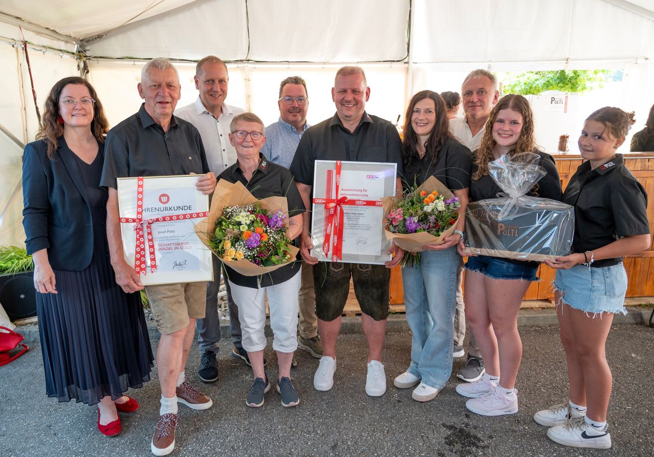 A group of people stands under a tent, holding flowers and certificates. A man and a woman hold a bouquet of flowers. Another man and a woman hold a certificate with a ribbon. Behind them, a man holds a box with a ribbon.