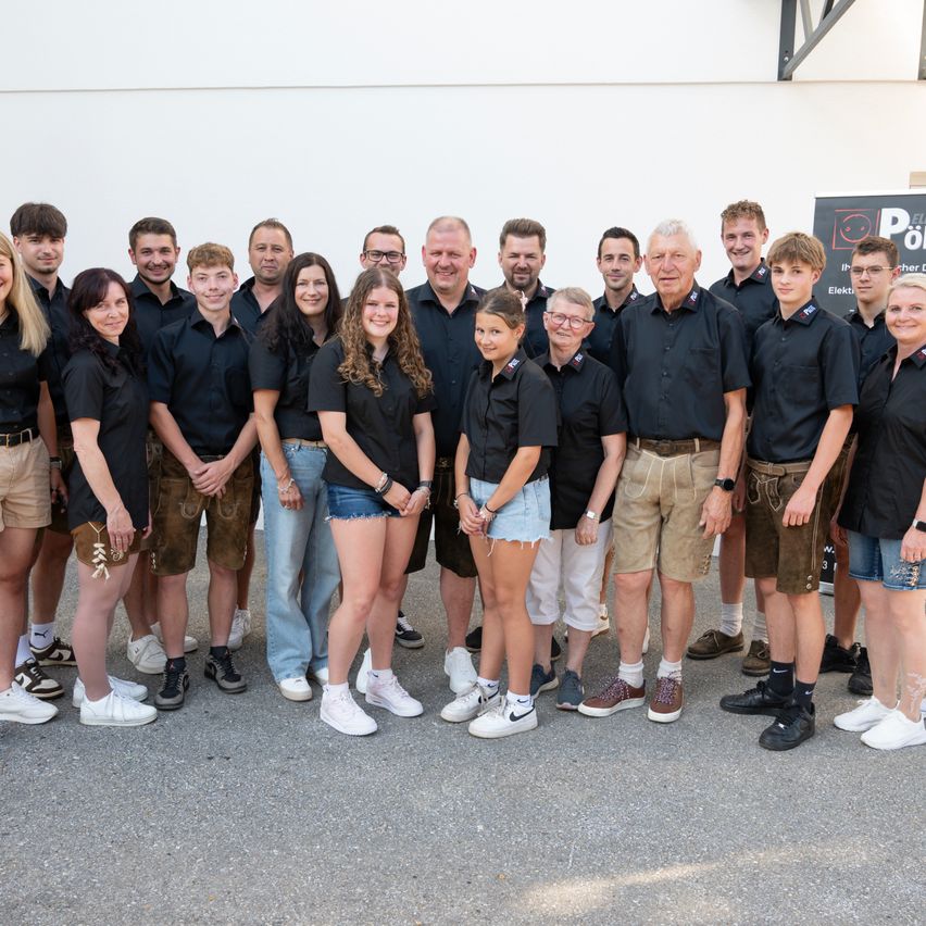 A group of individuals, likely employees, stand in a line wearing black shirts, shorts, and sneakers. Some are in traditional German attire. The setting appears to be outdoors.