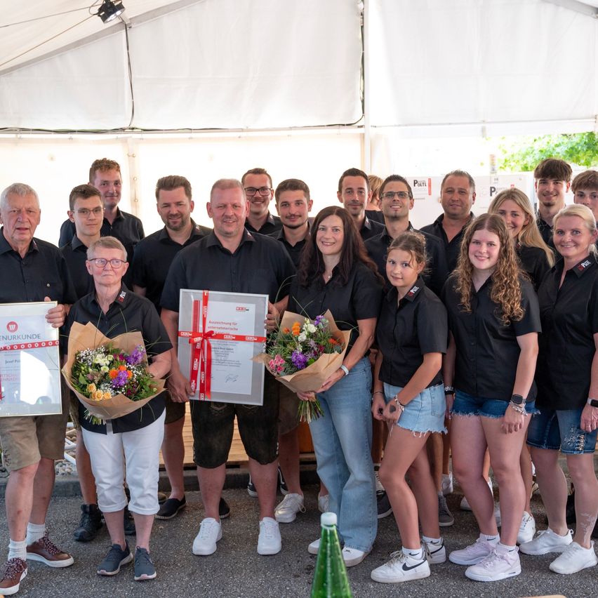 A group of people in black shirts and shorts stand under a tent, holding flowers and certificates. They appear to be celebrating a team achievement.