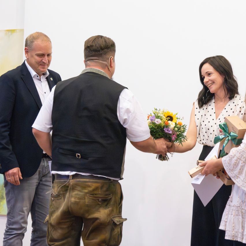Three people stand in a room; one man hands a bouquet of flowers to a woman, while a third individual holds a box with a ribbon.