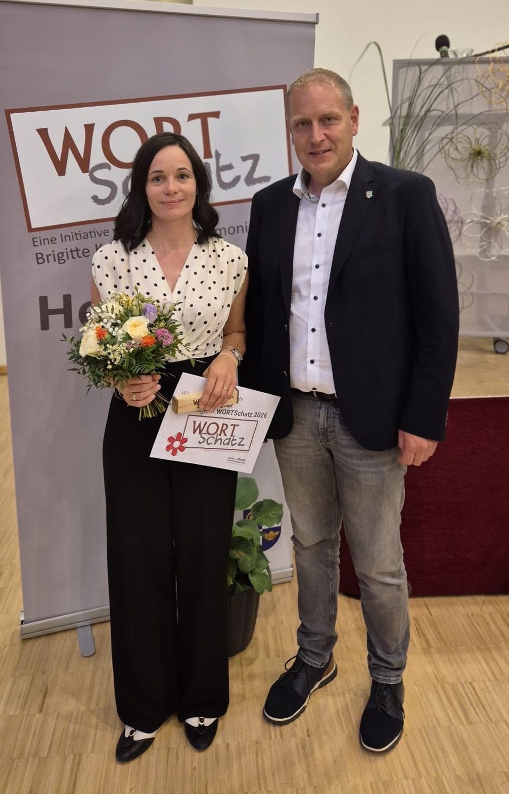 A woman holding flowers and a certificate stands beside a man, both smiling for a photo. Behind them is a banner with text and a plant.