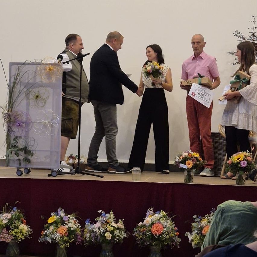 Five individuals stand on stage holding flowers and gifts, with a woman in a white blouse and black pants in the center. Two men stand on either side, one wearing a pink shirt, while two women stand at the ends, one wearing a white dress.