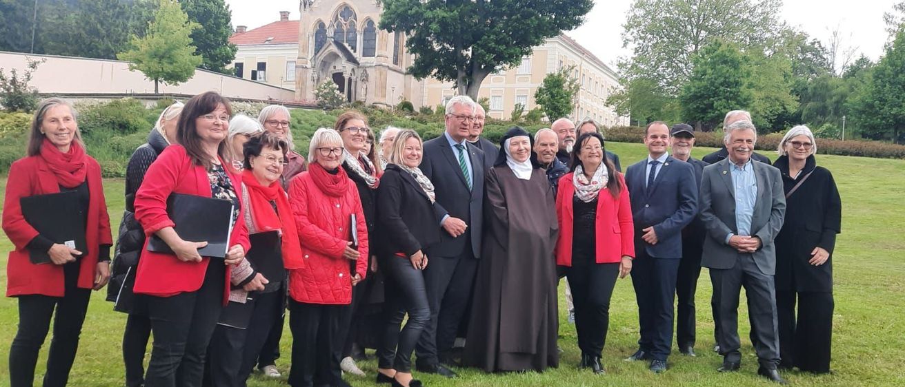 Eine Gruppe von Personen, einschließlich einer Nonne, steht in einem Park vor einem historischen Gebäude. Die Gruppe umfasst Menschen in Geschäftskleidung, Freizeitkleidung und Schals.