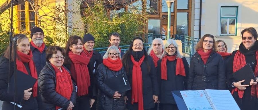 Ein Chor in roten Schals und schwarzen Mänteln steht vor einem Notenständer und einem Gebäude. Die Chormitglieder lächeln und scheinen sich auf eine Aufführung vorzubereiten.