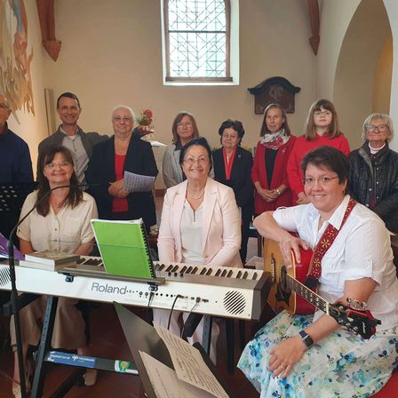 Eine Gruppe von Menschen steht in einer Kirche, einige lächeln und andere halten Bücher. Zwei Frauen spielen Klavier und Gitarre, mit einem Mikrofon und Notenblättern in der Nähe. Ein Buntglasfenster und ein Gemälde sind im Hintergrund zu sehen.