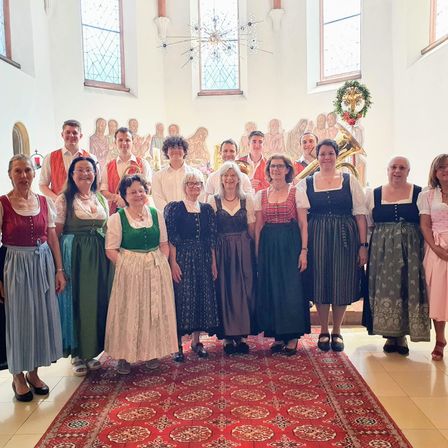 Eine Gruppe von Menschen in traditioneller Kleidung posiert für ein Foto in einer Kirche. Sie stehen vor einem Weihnachtskranz und Buntglasfenstern.