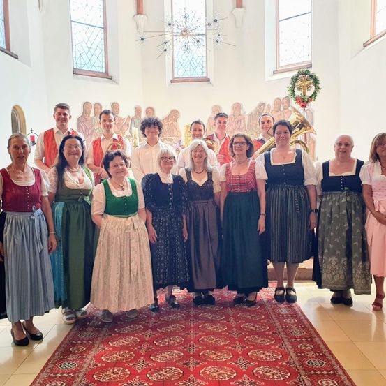Eine Gruppe von Menschen in traditioneller Kleidung posiert für ein Foto in einer Kirche. Sie stehen vor einem Weihnachtskranz und Buntglasfenstern.