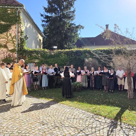 Ein Priester in einem gelben Gewand leitet einen Chor von Menschen in traditioneller Kleidung auf einem Kopfsteinpflaster-Vorhof mit einem Gebäude und Bäumen im Hintergrund.