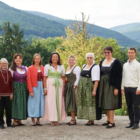 Eine Gruppe von Menschen, sowohl Männer als auch Frauen, in traditioneller Kleidung, steht vor einer Bergkette und Bäumen.