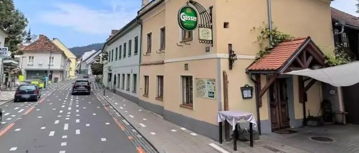 Ein Straßenblick zeigt ein Gebäude mit einem Schild, das 'Gusser' steht. Ein Tisch und ein Auto stehen vor dem Gebäude. Bäume und Gebäude befinden sich auf beiden Seiten der Straße.