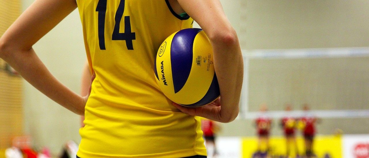 Eine Frau in einem gelben Trikot mit der Nummer 14 hält einen Volleyball. Der Hintergrund ist unscharf und zeigt andere Spieler auf dem Spielfeld.