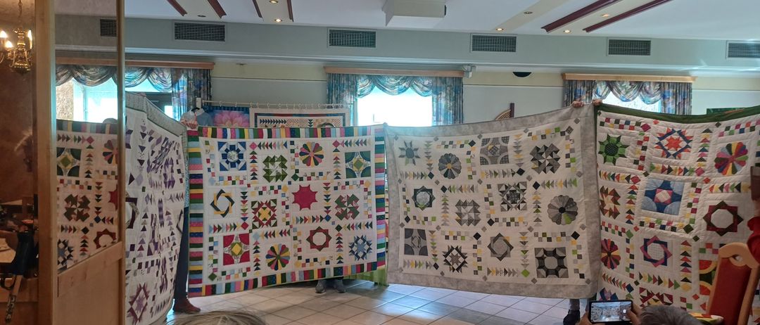 Eine Gruppe von Menschen präsentiert bunte Quilts in einem Raum mit gefliesten Böden. Die Quilts haben verschiedene Muster und Designs, und einige von ihnen haben ein Blumenmuster. Eine Person steht auf der linken Seite und eine andere Person steht auf der rechten Seite. Eine Person hält ein Telefon und scheint ein Foto zu machen.