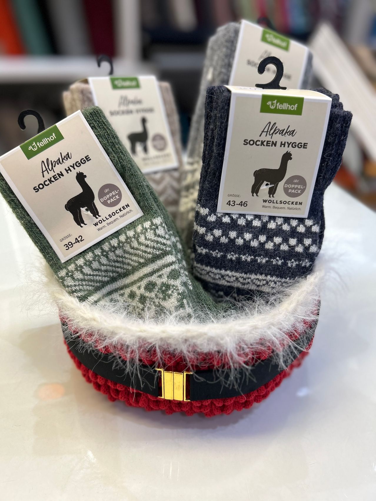Zwei Paar Alpaka-Socken sind in einem dekorativen rot-weißen Korb mit goldener Schnalle ausgestellt. Jedes Paar hat ein unterschiedliches Alpaka-Design und Etikett, eines in Grün und Weiß, das andere in Schwarz und Weiß.