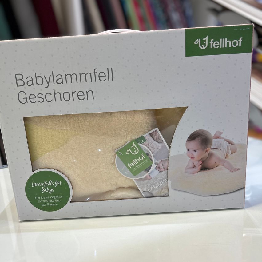 Eine Schachtel mit Fellhof Babylammwollfäden. Die Schachtel zeigt ein Bild eines Babys, das auf einer beigen Decke liegt, mit dem Text 'Babylammfell Geschenken'. Das Etikett enthält ein Babybild und Text auf Deutsch.