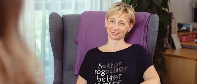 Eine Frau sitzt in einem Stuhl und trägt ein schwarzes T-Shirt mit der Aufschrift 'besser zusammen' und lächelt. Hinter ihr steht eine Pflanze und ein Schreibtisch.