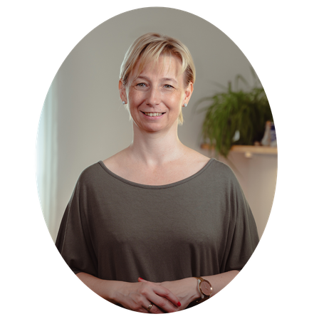 Eine Frau mit kurzen blonden Haaren lächelt und posiert für ein Foto. Sie trägt eine graue Bluse und hat eine Uhr am linken Handgelenk. Im Hintergrund steht eine Pflanze.