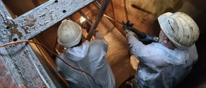 Zwei Personen in Schutzanzügen und Helmen arbeiten an einer Industriebaustelle, eine hält ein Werkzeug und die andere benutzt einen Bohrer.