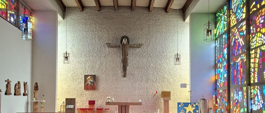 Ein leerer Kircheninnenraum mit Holzbänken, einem großen Kreuz an der Wand und farbenfrohen Buntglasfenstern. Der Altar ist mit Blumen geschmückt, und ein roter Teppich führt dazu.