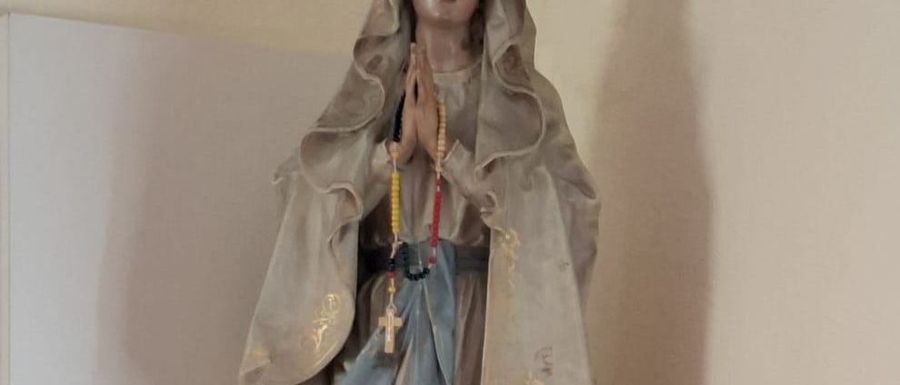 Eine Statue einer betenden Frau steht auf einem weißen Regal, trägt ein blaues Gewand und hält einen Rosenkranz. Zwei rote Votivkerzen befinden sich auf beiden Seiten der Statue.