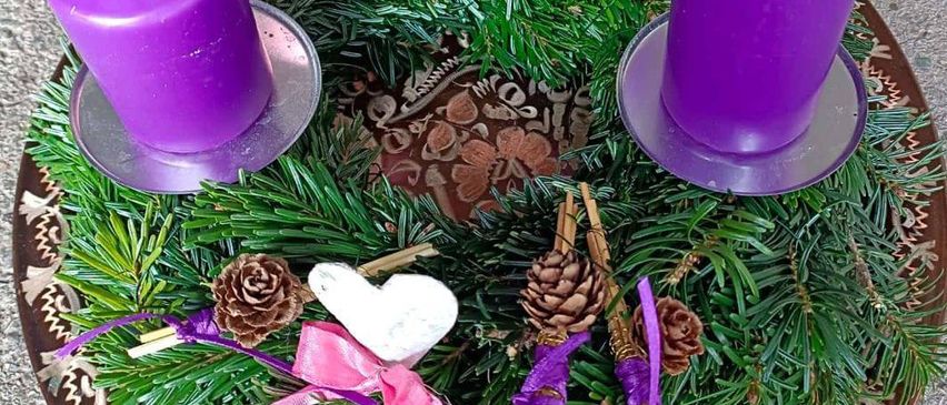 Ein Kranz mit vier lila Kerzen, eine davon rosa, steht auf einem Tisch. Grünes Laub und Tannenzapfen schmücken den Kranz. Ein Textüberlagerung lautet 'ADVENTSINGEN' und die Veranstaltungsdetails.