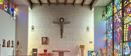 Das Innere einer Kirche zeigt ein großes Kreuz an einer weißen Wand, Reihen von Kirchenbänken und Buntglasfenster. Ein roter Teppich führt zu einem mit Blumen geschmückten Altar.
