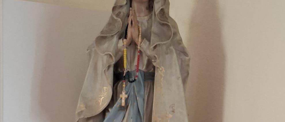 Eine Statue einer betenden Figur mit einem Rosenkranz, die ein blaues Gewand trägt, steht auf einem weißen Regal mit zwei roten Kerzen.