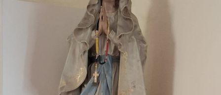 Eine Statue einer betenden Figur mit einem Rosenkranz, die ein blaues Gewand trägt, steht auf einem weißen Regal mit zwei roten Kerzen.