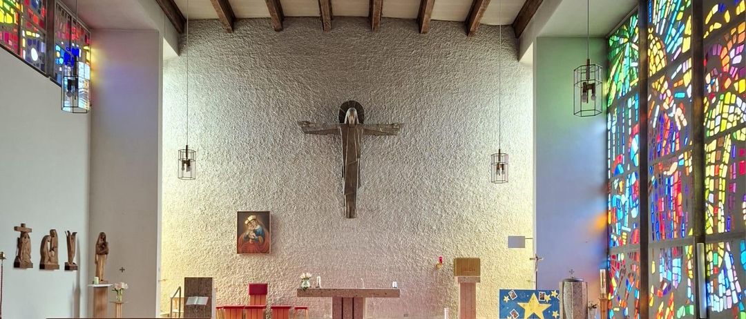Eine leere Kirche mit Holzbänken, einem roten Teppich und einem Kreuz an der Wand. Es gibt Buntglasfenster und Lampen, die von der Decke hängen.