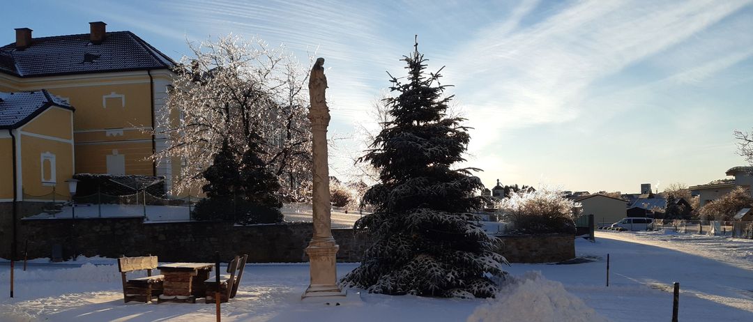 Eine Statue steht auf einem Podest, umgeben von Schnee. Ein Tannenbaum steht neben ihr, ebenfalls mit Schnee bedeckt. Dahinter befindet sich ein Gebäude mit einer Ziegelmauer. Der Himmel ist blau mit einigen Wolken.