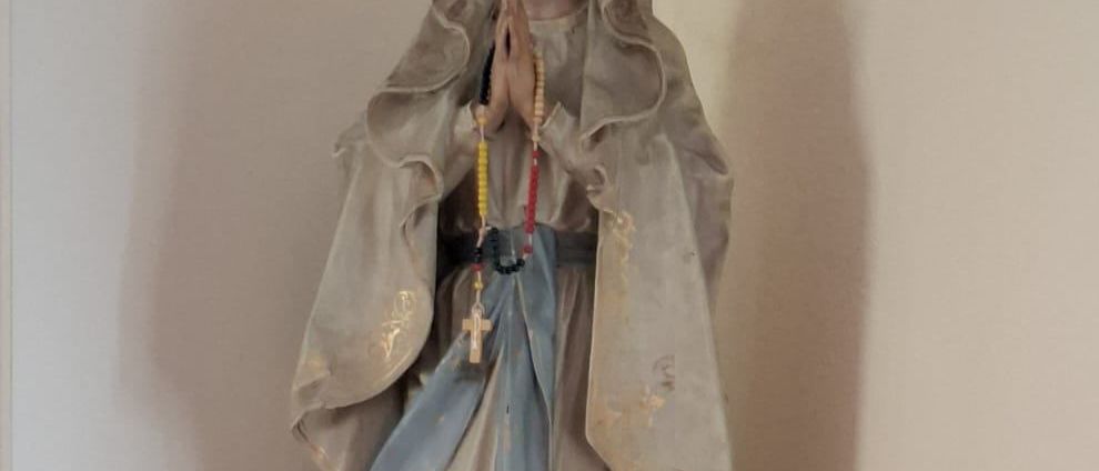 Eine Statue einer Heiligen mit gefalteten Händen in Gebetshaltung. Eine Rosenkranzkette befindet sich um ihren Hals. Zwei rote Kerzen stehen auf beiden Seiten der Statue.