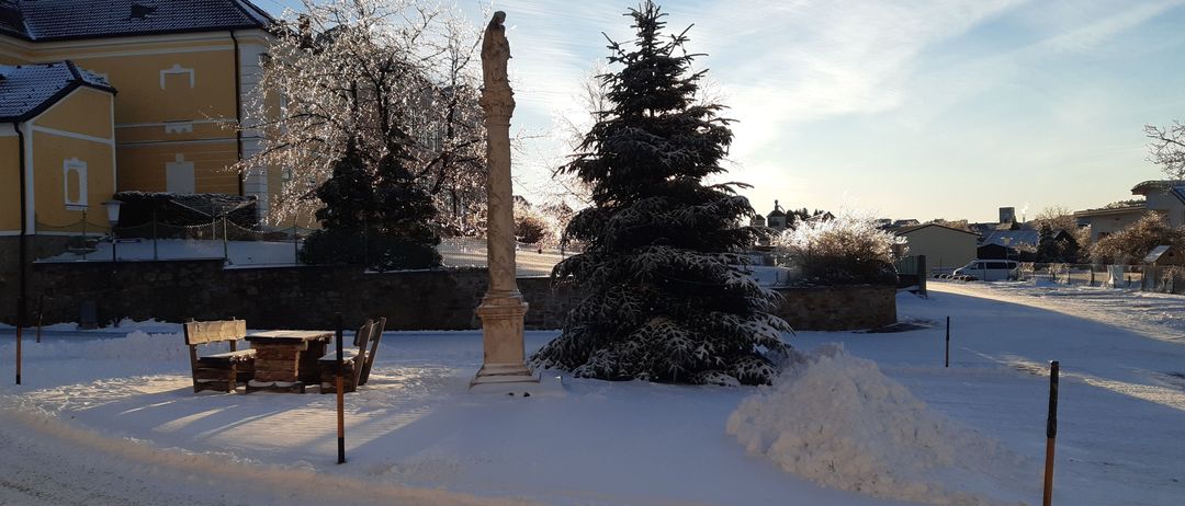 Ein schneebedeckter Park zeigt eine Statue auf einer Säule und einen großen, schneebedeckten Tannenbaum. Ein Holzstuhl und ein kleiner Tisch sind ebenfalls vorhanden.