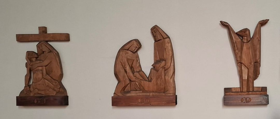 Eine Holzschnitzerei, die zwei Figuren darstellt, eine kniend und die andere stehend, an einer weissen Wand befestigt. Die Schnitzerei hat ein Etikett mit der Aufschrift 'XXM'.