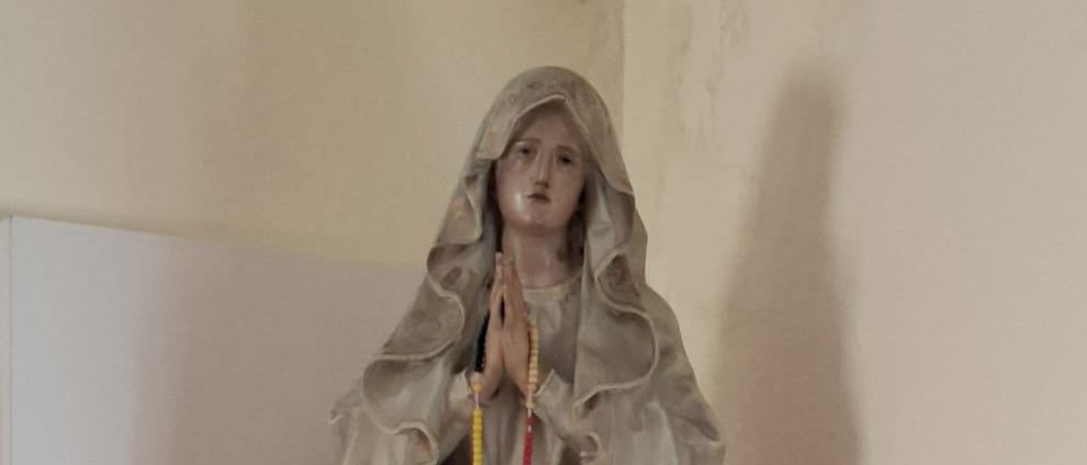 Eine Statue von Maria mit einem Rosenkranz steht auf einem weißen Regal, mit zwei roten Kerzen auf beiden Seiten.