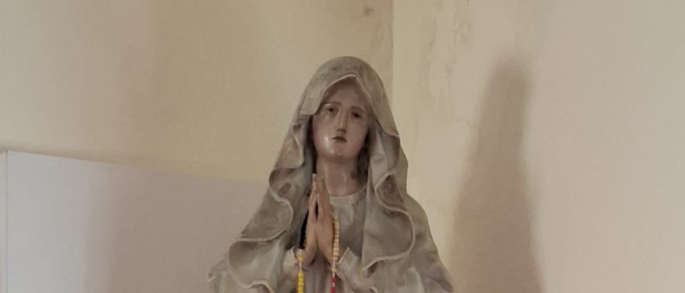 Eine Statue einer Frau mit einem Schleier steht auf einem weißen Regal. Sie hält ein Rosenkranz und ein Kreuz. Zwei rote Kerzen stehen neben ihr.