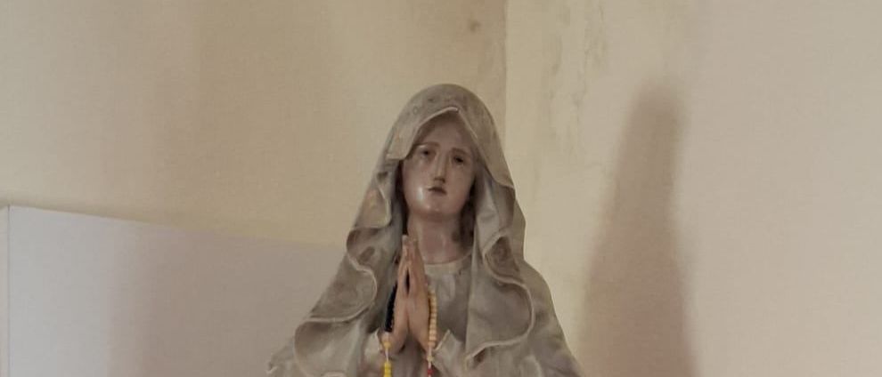 Eine Statue einer betenden Frau mit einem Rosenkranz, auf einem weißen Regal mit zwei roten Kerzen platziert.