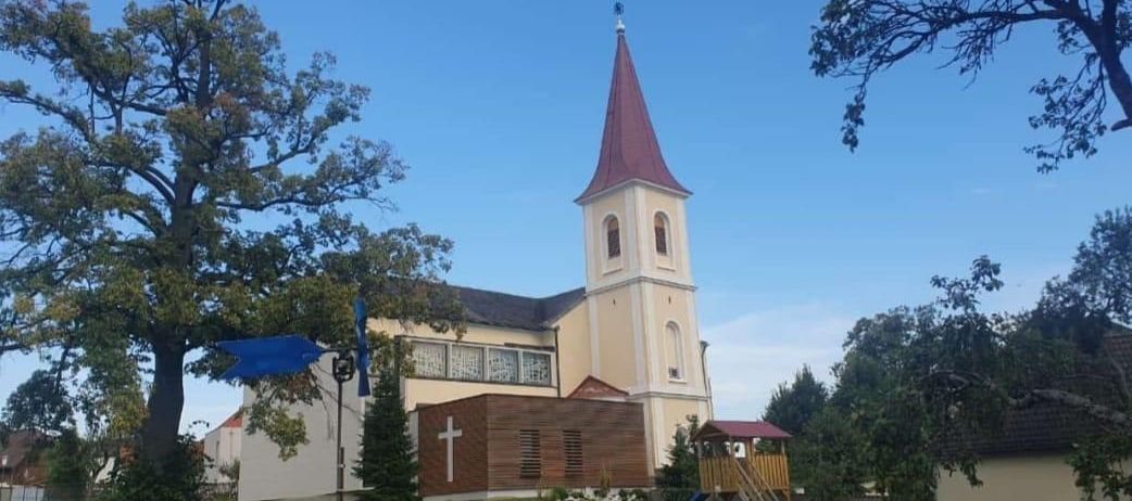 Eine beigefarbene Kirche mit einem Kirchturm und einem Kreuz an der Wand. Eine blaue Flagge steht davor. Ein Baum steht vor der Kirche. Ein Spielplatz befindet sich hinter der Kirche.
